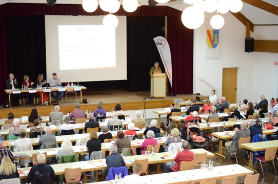 Blick von oben auf den Versammlungssaal mit besetzten Tischreihen, Bühne und Rednerpult.