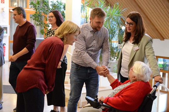 Mehrere Menschen begegnen sich im Foyer; ein Mann begrüßt eine ältere Frau im Rollstuhl mit Handschlag.
