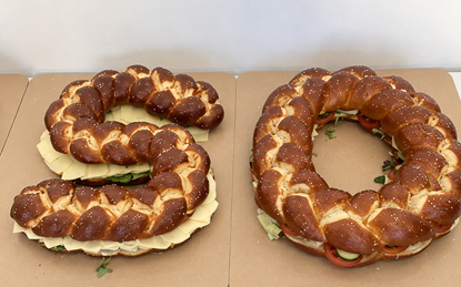 Zwei große, mit Käse, Salat und Gemüse belegte Laugenbrezeln liegen auf einer Tischfläche. Die linke Brezel hat die Form einer Fünf, die rechte die Form einer Null.