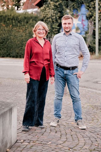 Karen Eßrich in roter Bluse und Oliver Hill in gemustertem Hemd stehen nebeneinander auf einem gepflasterten Platz.