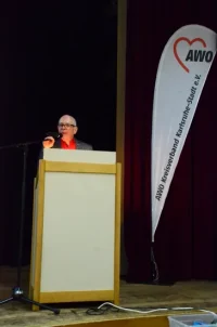 Gerolf Heberling steht mit einem Mikrofon am Rednerpult. Rechts ist ein AWO-Banner zu sehen.