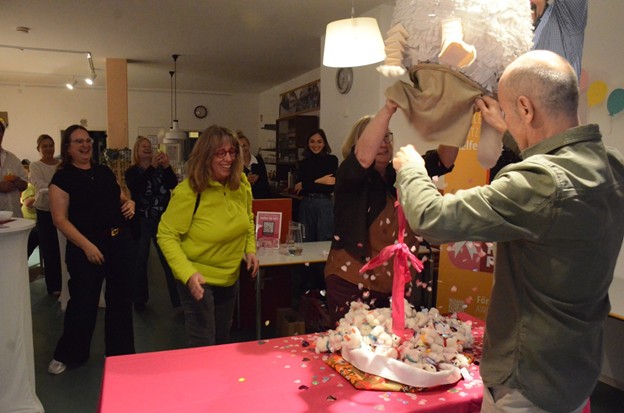 Die Hunde-Piñata wird angehoben. Darunter liegen kleine Plüschhunde auf einem Tisch, Konfetti fällt herab und die Gäste lachen.