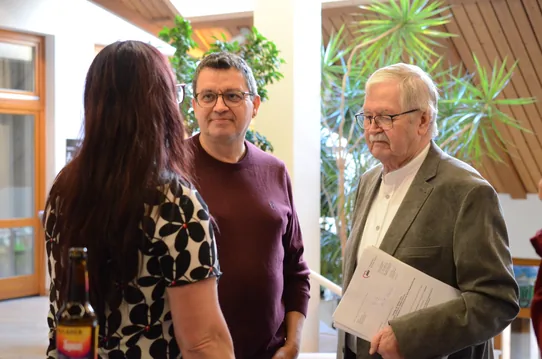 Drei Personen stehen im Foyer im Gespräch. Der ältere Mann rechts hält ein Dokument des AWO Kreisverbands Karlsruhe-Stadt.