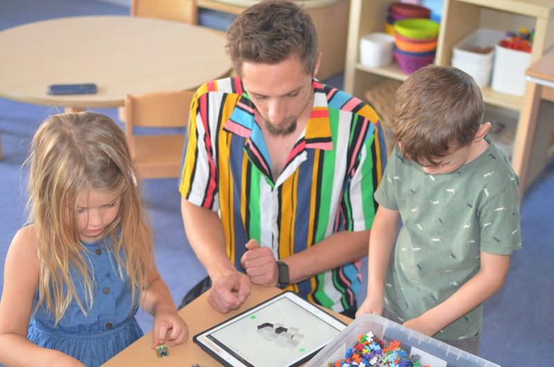 Ein Erzieher und zwei Kinder schauen gemeinsam auf ein Tablet. Auf dem Tisch liegen bunte Steckbausteine.