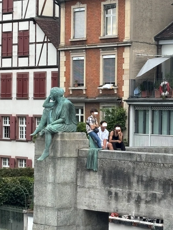 Drei Menschen sitzen neben einer großen sitzenden Bronzestatue auf einem steinernen Sockel.